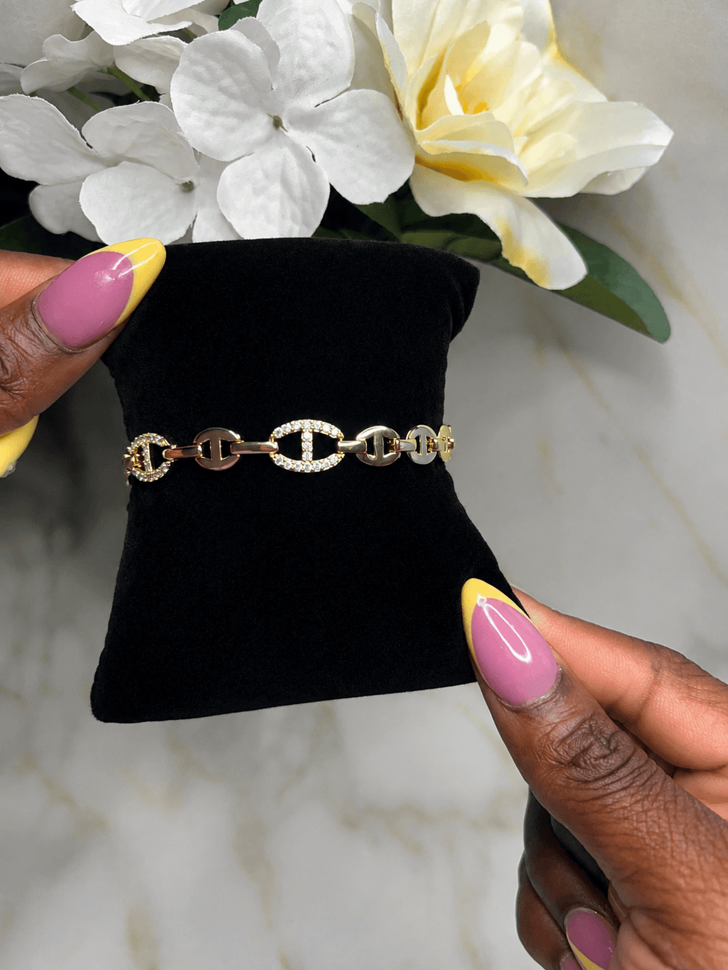 A gold chain bracelet with interlocking oval links, some adorned with small clear stones, displayed on a black velvet jewelry stand held by two hands with pink and yellow manicured nails, with white flowers in the background.
