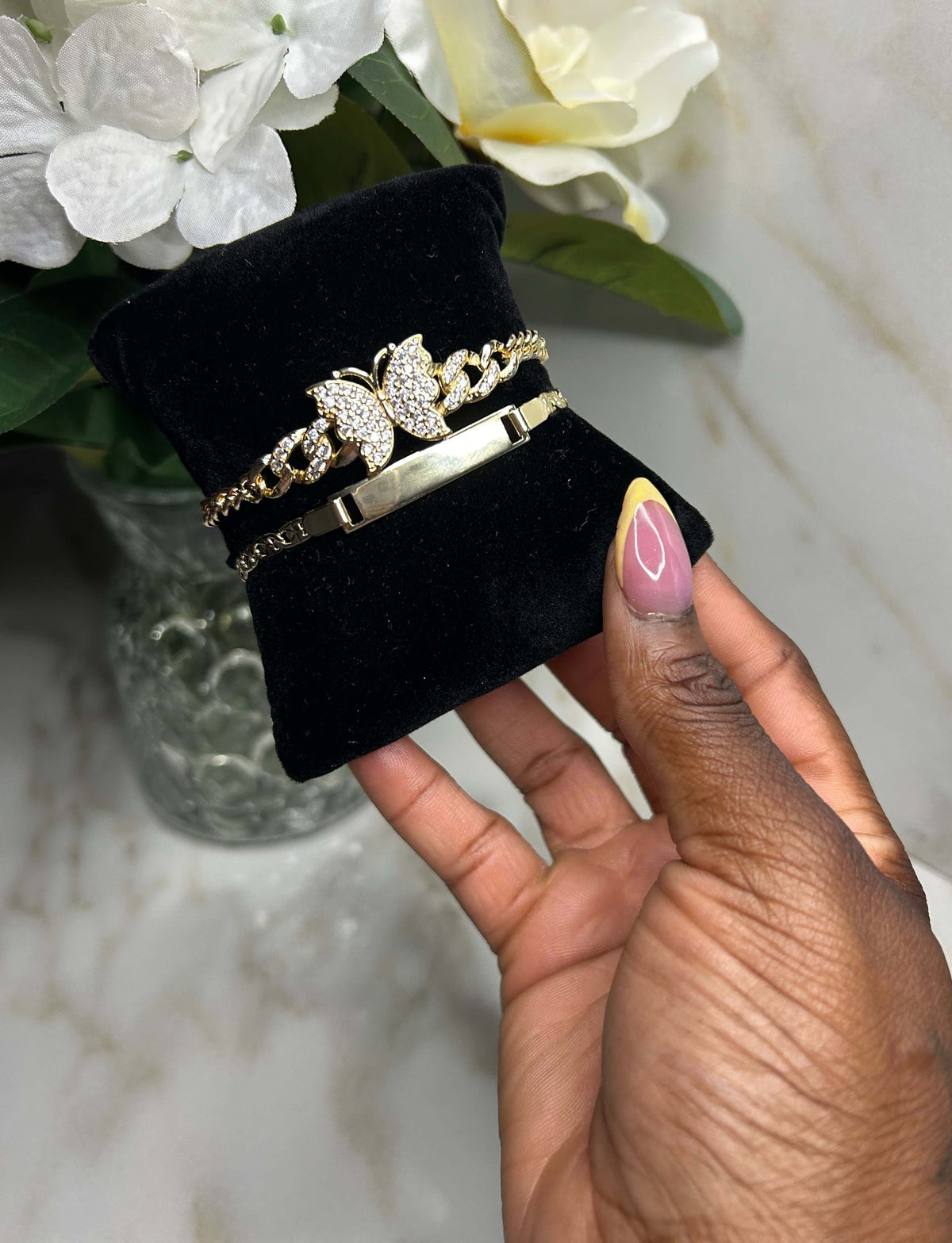 A hand with pink and yellow nail polish holds a black velvet jewelry display cushion featuring three gold bracelets, including a butterfly design with clear rhinestones and a polished bar bracelet, against a marble background with white flowers.