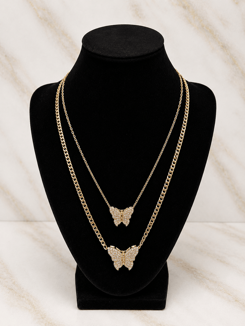Two layered gold-tone chain necklaces with butterfly pendants, displayed on a black velvet jewelry bust against a light marble-patterned background.