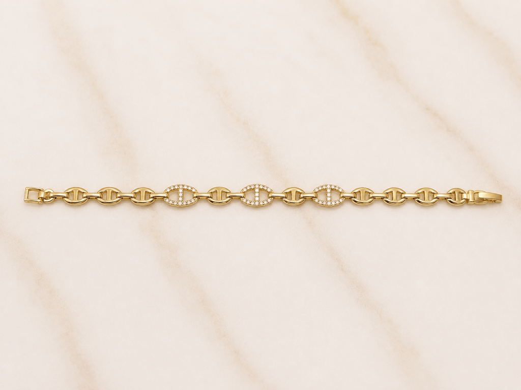 A gold chain bracelet with three circular links set with small clear stones, displayed horizontally on a light beige marble surface.