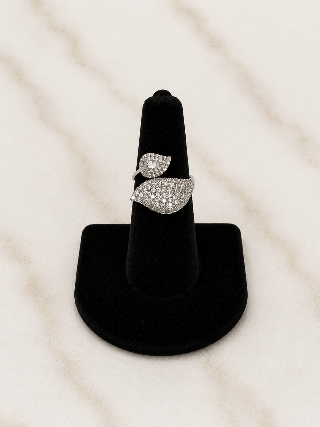 A silver ring with pavé-set clear stones in a leaf-like design, displayed on a black ring stand against a light marble background.