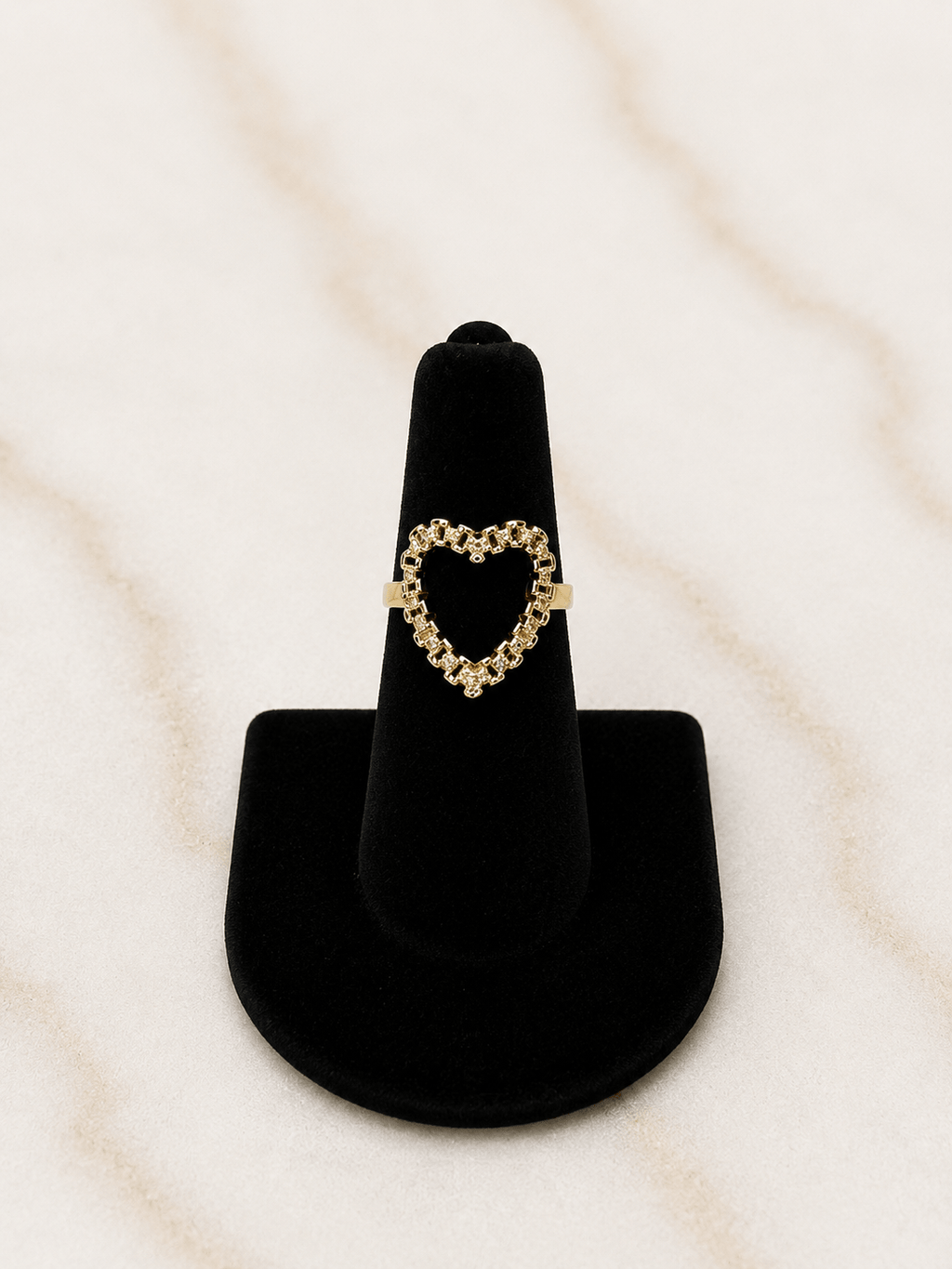 A gold heart-shaped ring displayed on a black velvet ring stand against a light beige marble background.