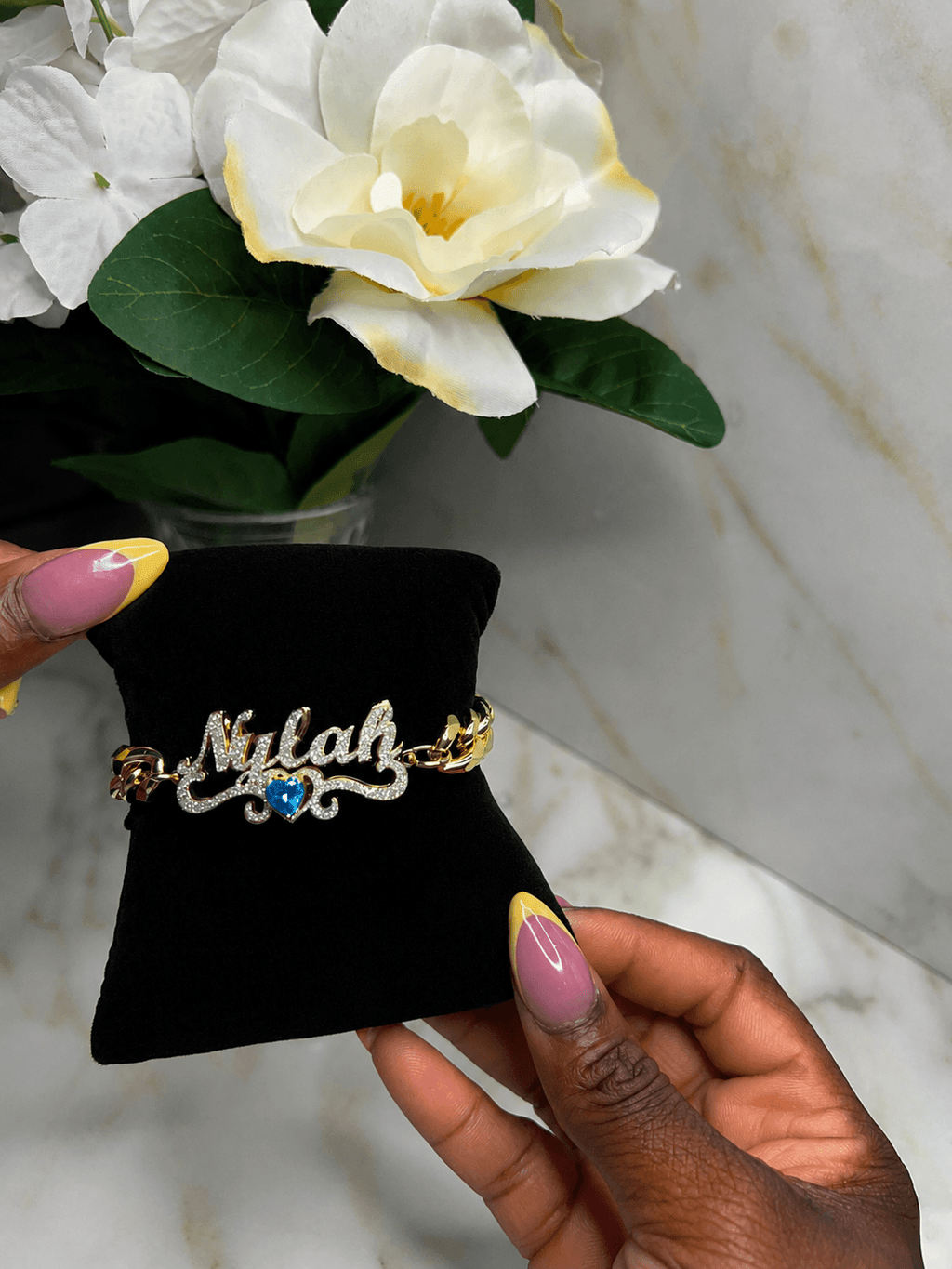 A close-up of a hand holding a black velvet jewelry display pad with a nameplate bracelet that reads 'Nilah' in silver script, accented with gold chain links and a small blue heart. The background shows a marble surface and white flowers.