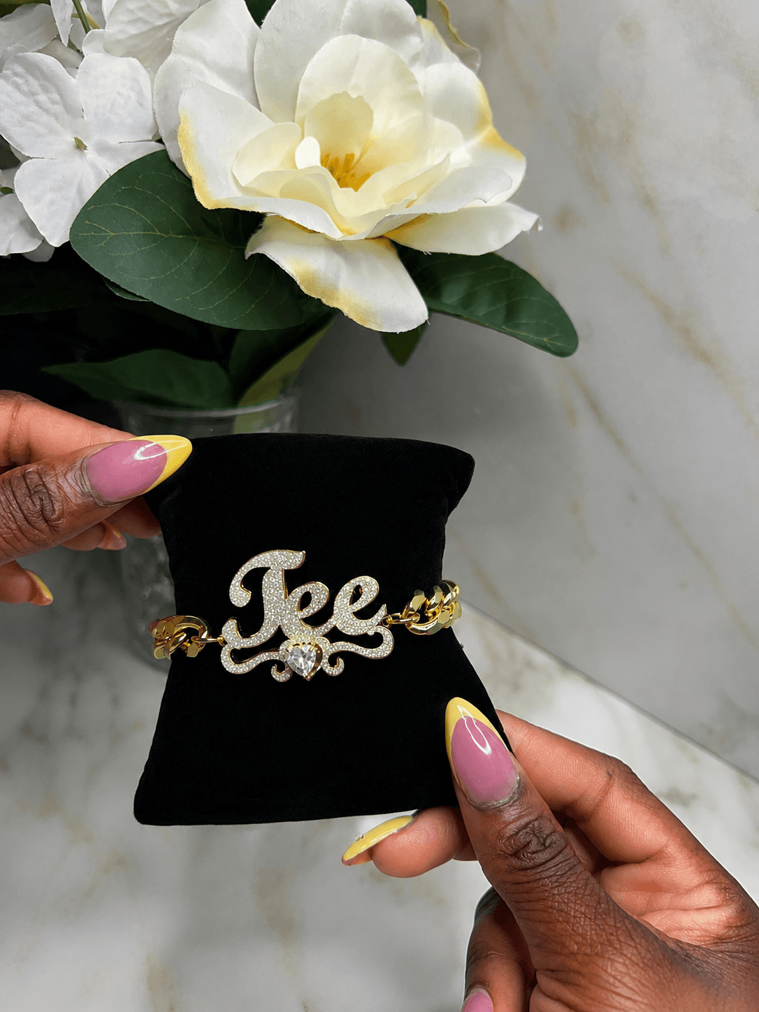 A gold and crystal bracelet with the word 'Jee' in cursive lettering, displayed on a black velvet jewelry stand held by hands with pink and yellow manicured nails, with white flowers in the background.