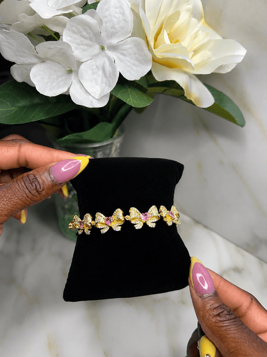 A gold-toned bracelet with multiple bow motifs set with pink gemstones and clear crystals, displayed on a black velvet jewelry stand held by hands with pink and yellow nail polish, with white flowers in the background.
