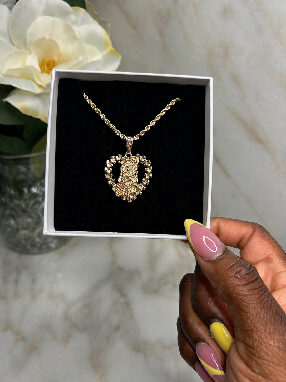 A gold heart-shaped pendant necklace with a central figure, displayed inside a white gift box with a black velvet lining. A hand with pink and yellow manicured nails holds the box, set against a marble background with a white flower in a vase.