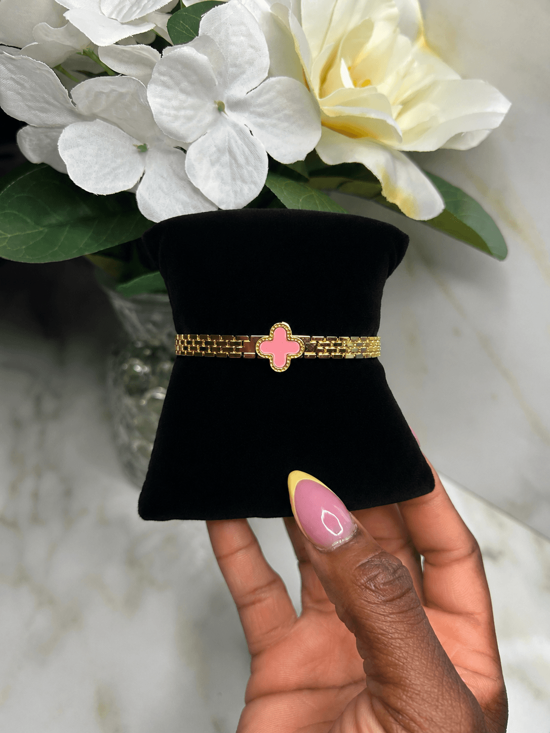A gold beaded bracelet with a pink enamel clover charm, displayed on a black velvet jewelry stand held by a hand with pink and yellow nail polish, with white flowers in the background.