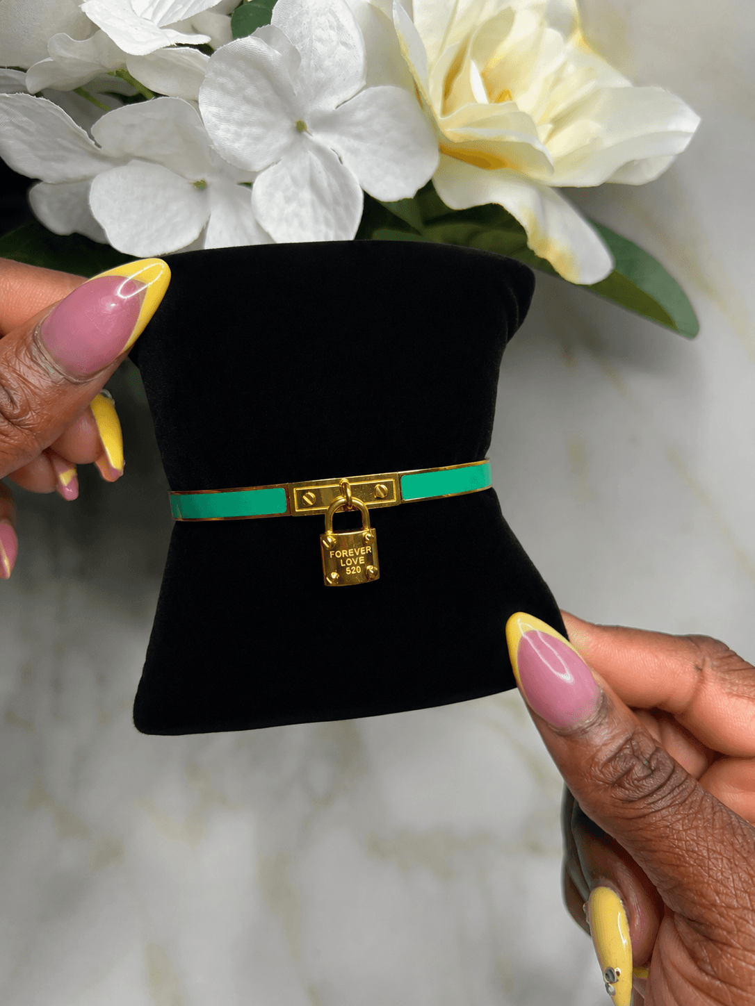 A green and gold faux-leather bracelet with a gold padlock charm, displayed on a black velvet miniature top hat stand held by hands with pink and yellow manicured nails, with white flowers in the background.