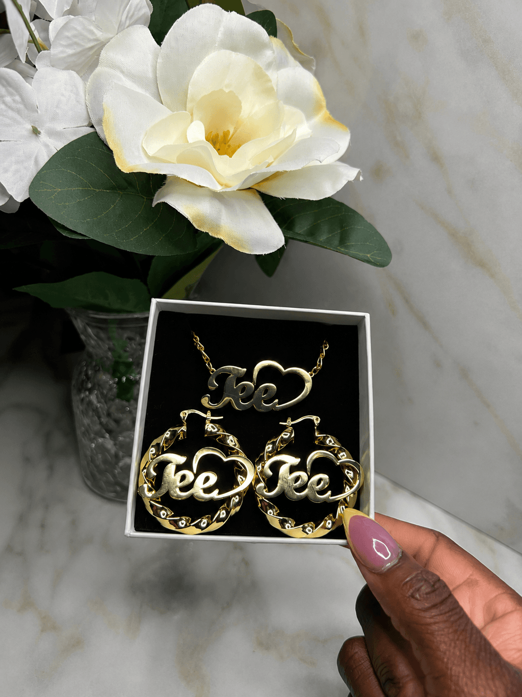 A hand with pink nail polish holds an open white jewelry box containing a gold-tone necklace and matching hoop earrings, each with a cloud-shaped center and the word 'Tee' in script. White flowers in a vase sit in the background on a light marble surface.