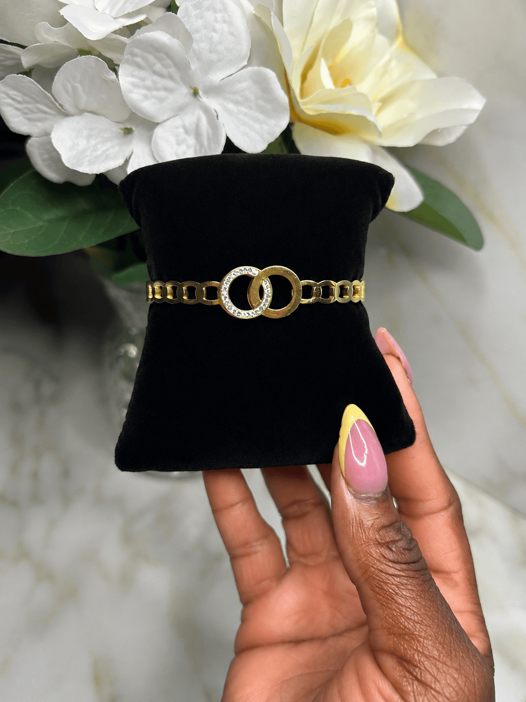 A gold chain bracelet with a double interlocking ring centerpiece, held by a hand over a black velvet jewelry display. White flowers are in the background.