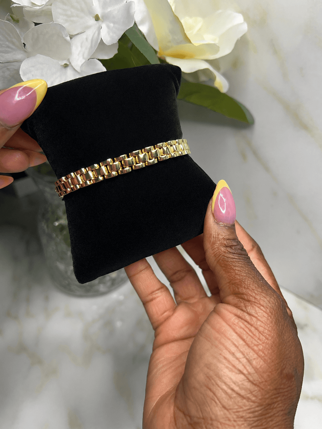 A gold chain link bracelet displayed on a black velvet jewelry stand, held by two hands with pink and yellow manicured nails against a marble background with white flowers.