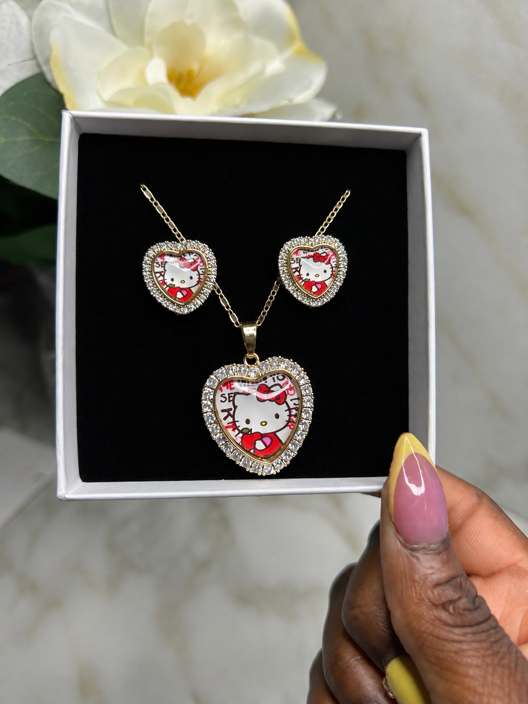 A gold heart-shaped jewelry set featuring a pendant necklace and matching earrings with a Hello Kitty design, displayed in a white gift box with a black insert. A hand with pink and yellow nail polish holds the box, with a white flower and a light marble background visible.