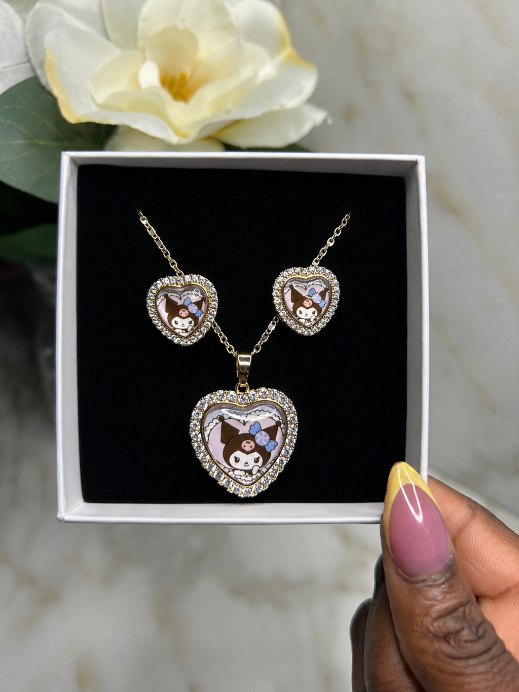 A heart-shaped pendant necklace and matching heart-shaped earrings displayed in an open white gift box with a black insert. The jewelry has a gold-tone outline, clear rhinestone borders, and cartoon character enamel details.
