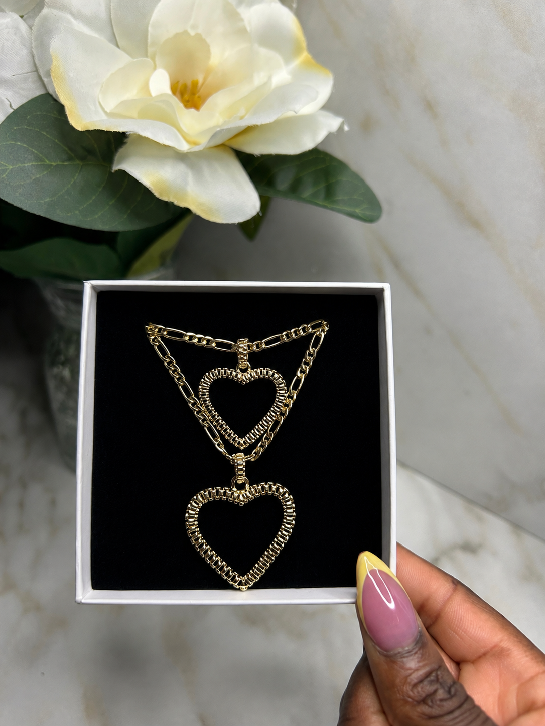 A gold heart pendant necklace displayed inside a black jewelry box, held by a hand with pink and yellow nail polish, with a white flower and marble background.