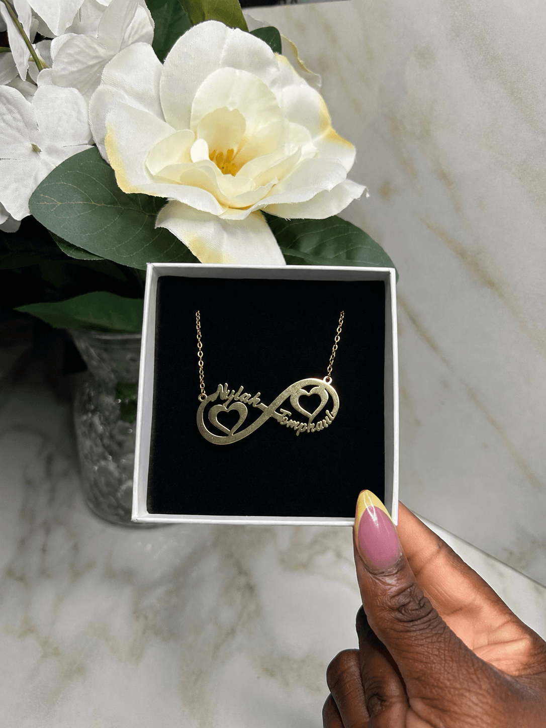 A gold infinity necklace with two interlocking heart shapes and the words 'Love Family' engraved, presented in a black box and held by a hand against a marble background with white flowers.