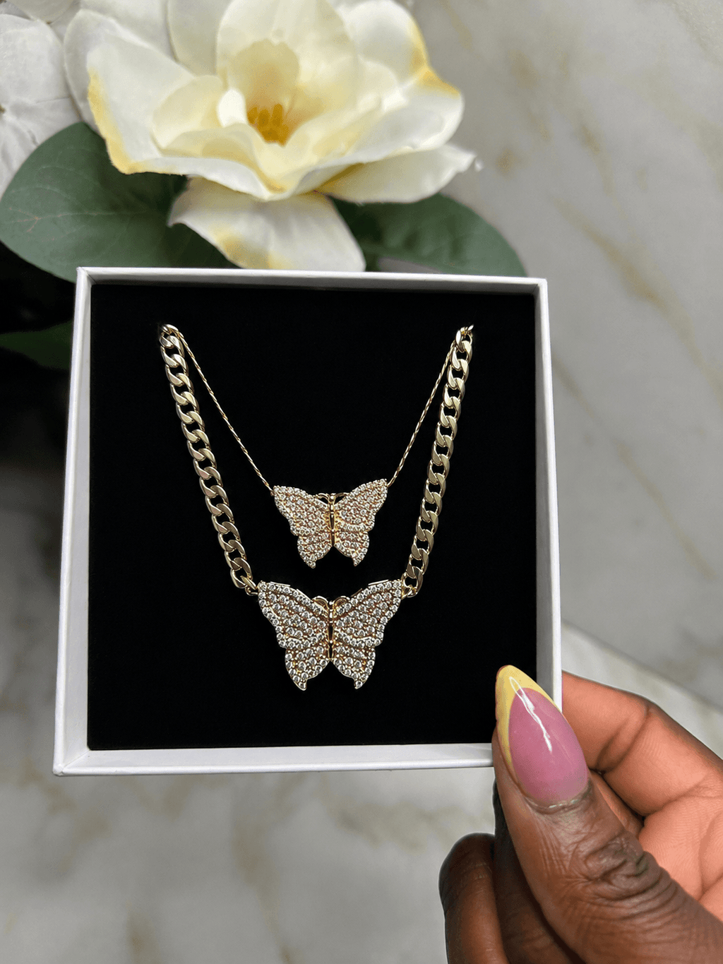 A gold-tone double-layered necklace with two butterfly pendants covered in clear rhinestones, presented in a white box with a black velvet insert and held by a hand with pink and yellow manicured nails.