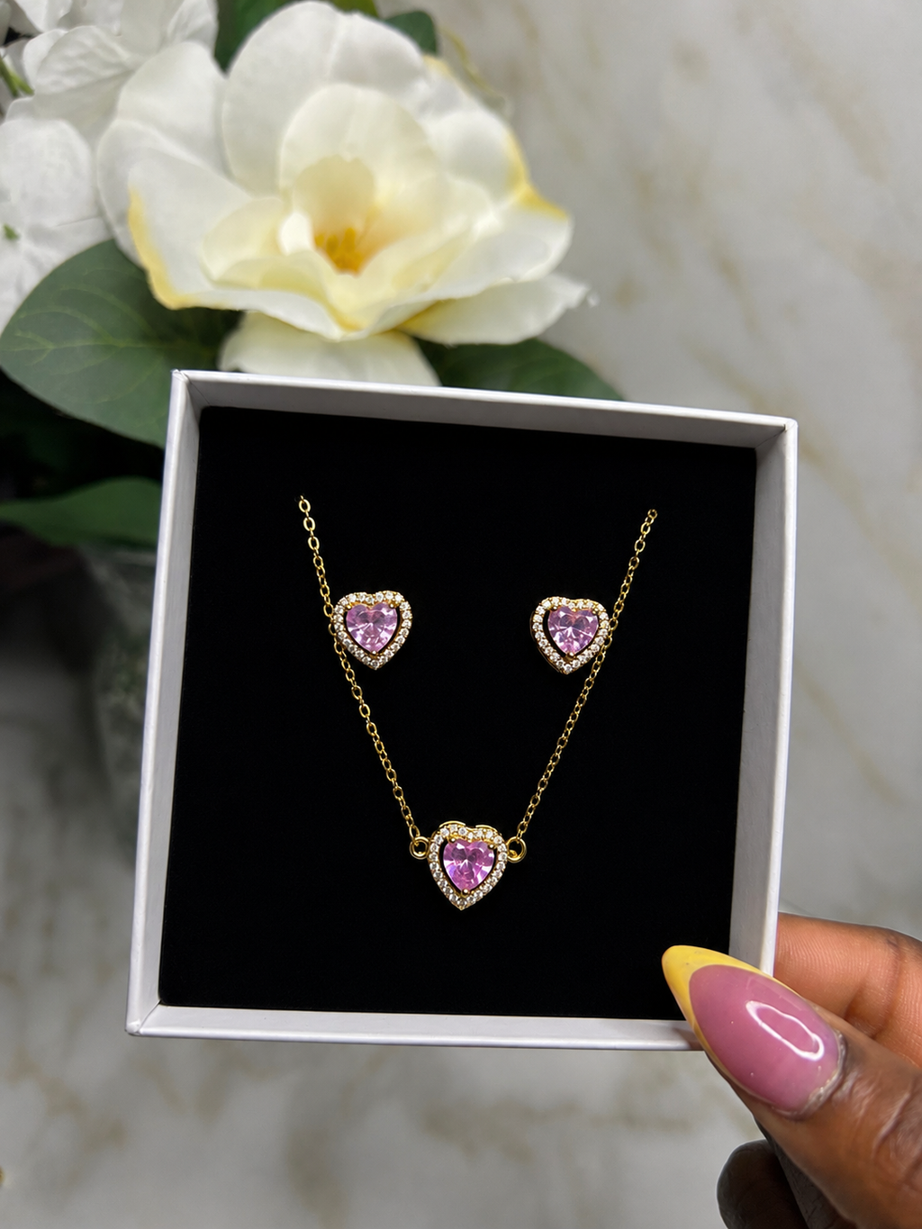 A gold necklace and matching stud earrings with heart-shaped pink gemstones, presented in a white gift box with a black insert. A hand with pink and yellow nail polish holds the box, with a white flower and marble surface in the background.