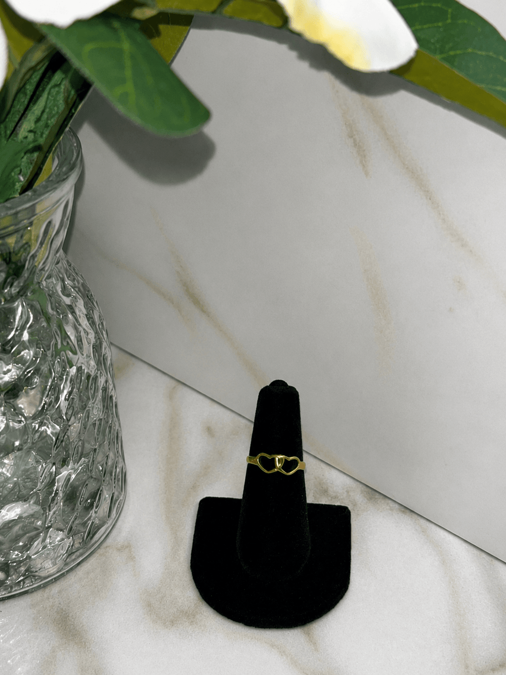 The Golden Bond Ring displayed on a black stand next to a glass vase, showcasing its elegant double heart design.