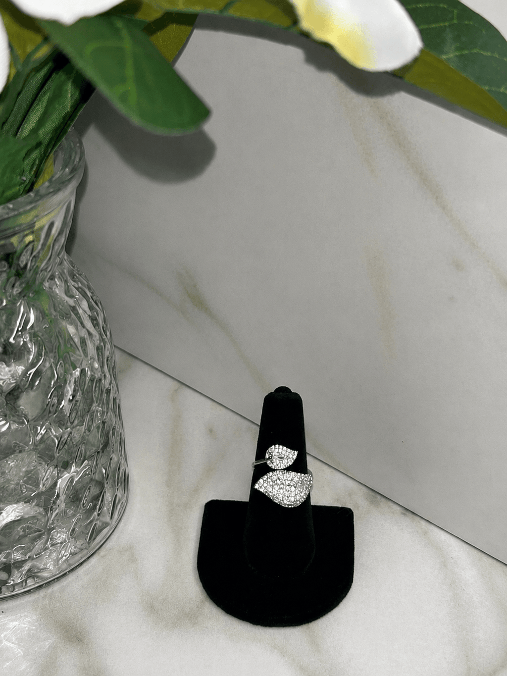 A silver ring with a leaf-shaped pavé setting displayed on a black ring stand, set against a marble background with a glass vase of flowers.