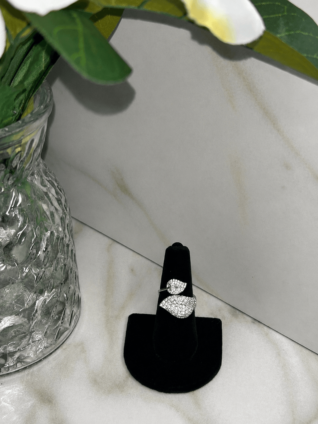 A silver ring with a leaf-shaped pavé setting displayed on a black ring stand, set against a marble background with a glass vase of flowers.