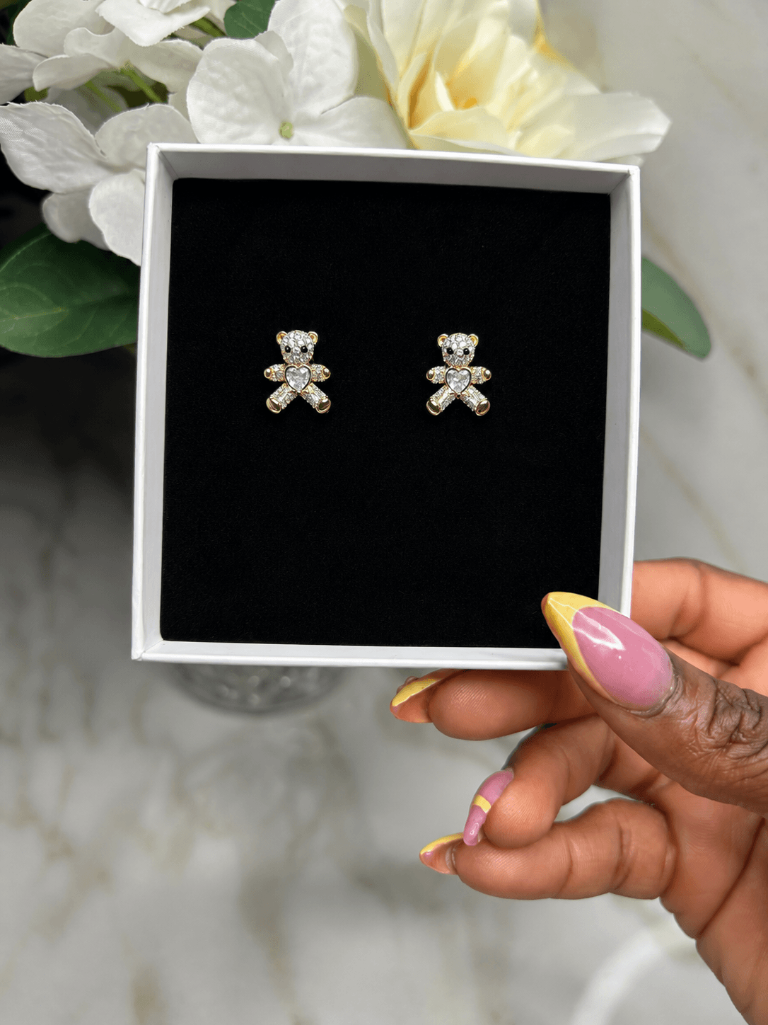 A pair of gold teddy bear stud earrings with clear rhinestones, presented in a white gift box with a black velvet insert, held by a hand with pink and yellow manicured nails.
