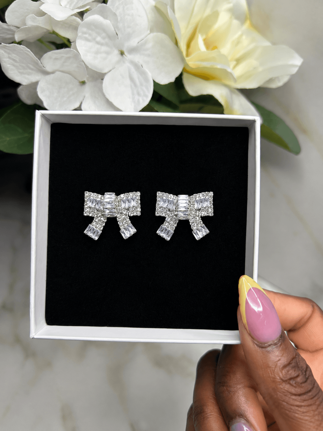 A pair of silver bow-shaped stud earrings covered in clear crystals, presented in a white gift box with a black velvet insert. A hand with pink and yellow manicured nails holds the box, with white flowers in the background.