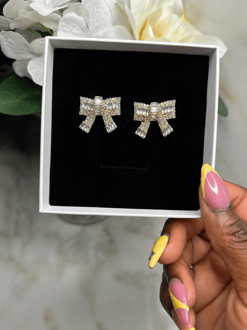A pair of silver bow-shaped stud earrings encrusted with clear crystals, presented in a white gift box with a black velvet insert, held by a hand with pink and yellow manicured nails.