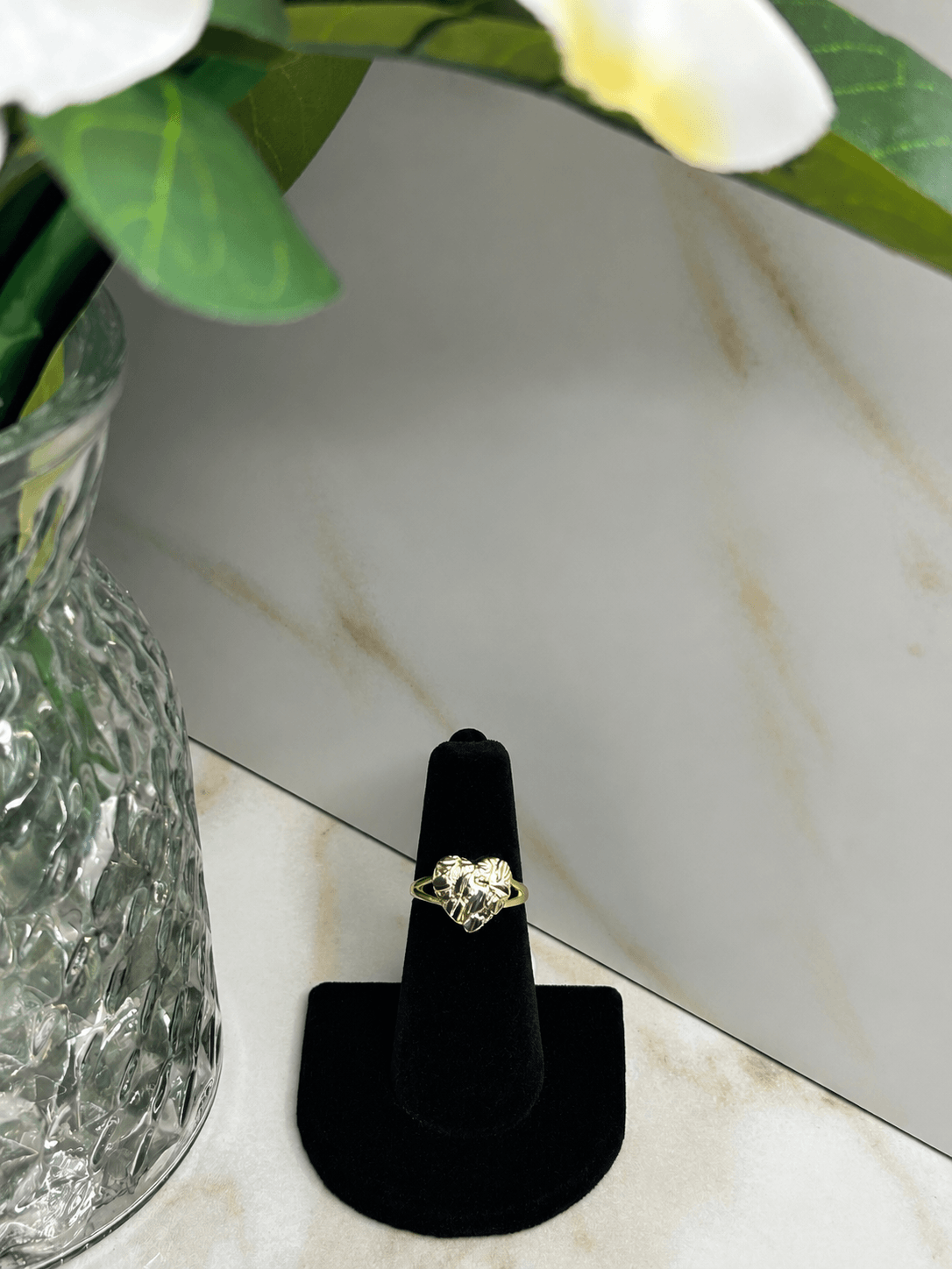 The Reign Golden Heart Ring displayed on a black velvet stand next to a flower vase, showcasing its elegant design.