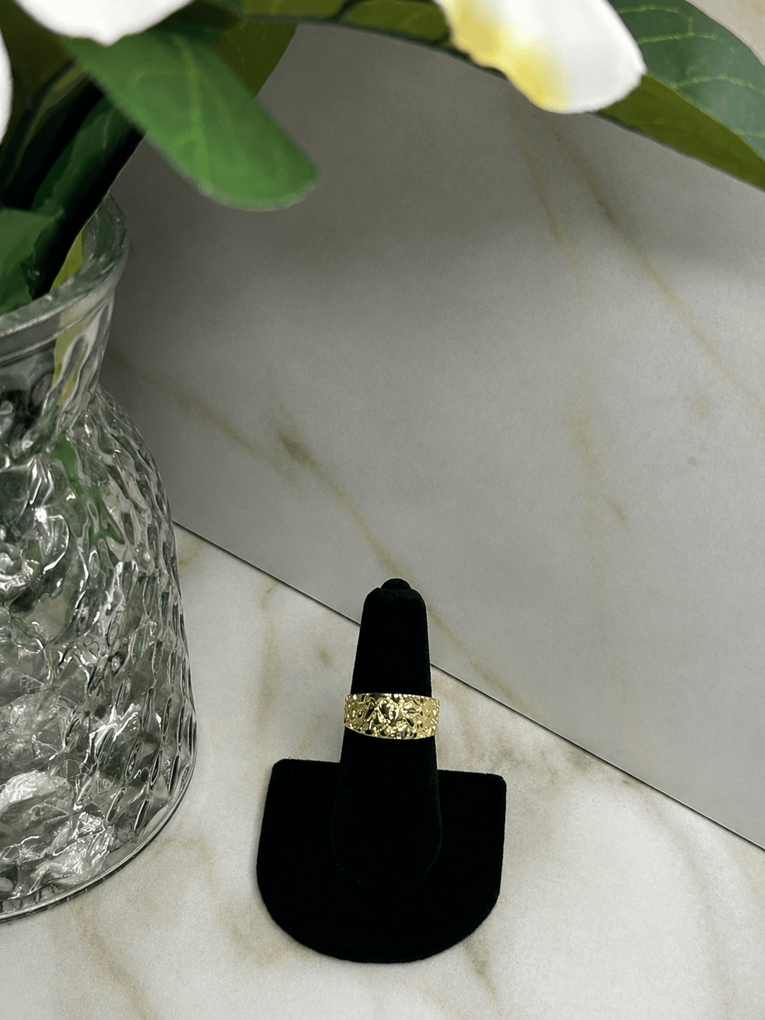 A gold ring with a textured, patterned band displayed on a black ring stand, set on a light marble surface next to a glass vase with flowers.