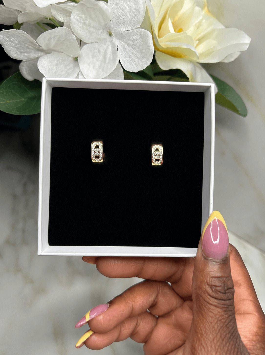 A pair of gold-toned rectangular stud earrings with clear stones, displayed on a black insert inside an open white gift box, held by a hand with pink and yellow manicured nails.