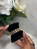 A gold double-strand bracelet with small clear rhinestone accents is displayed on a black velvet jewelry holder, held by two hands with pink and yellow manicured nails. The background features a white marble surface and a vase of white flowers.
