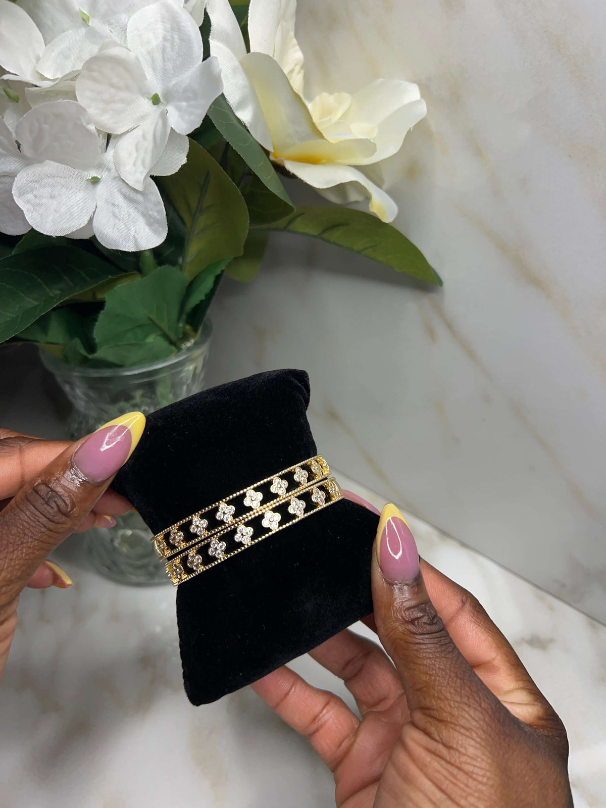 A gold double-strand bracelet with small clear rhinestone accents is displayed on a black velvet jewelry holder, held by two hands with pink and yellow manicured nails. The background features a white marble surface and a vase of white flowers.