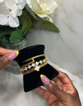 A close-up of a person's hands holding a black velvet jewelry display stand with three gold bracelets. The bracelets include a simple gold bangle, a gold chain with clear stones, and a gold chain with a white clover motif.