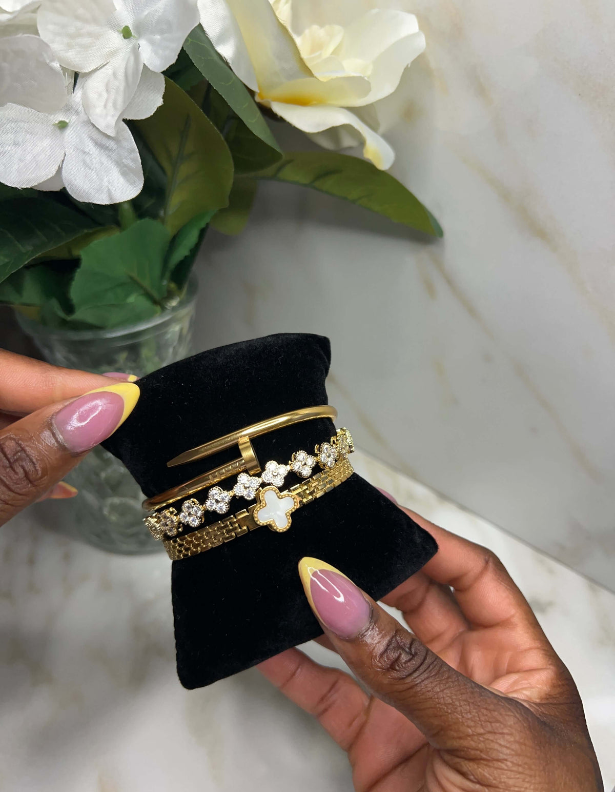 A close-up of a person's hands holding a black velvet jewelry display stand with three gold bracelets. The bracelets include a simple gold bangle, a gold chain with clear stones, and a gold chain with a white clover motif.