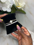 A close-up of a person's hands holding a black velvet jewelry display cushion with three delicate bracelets. The bracelets include a gold chain with a white mother-of-pearl four-leaf clover charm, a thin gold chain, and a silver chain with small clear stones.
