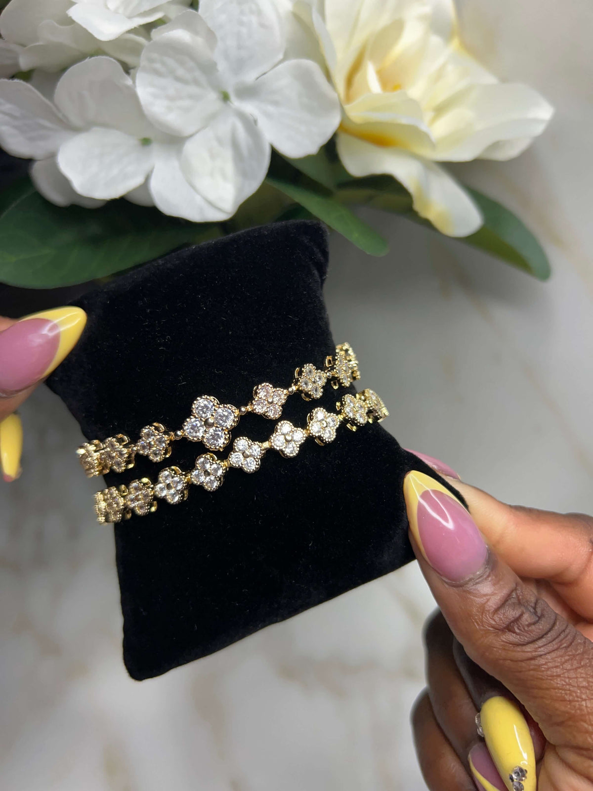 A pair of gold-tone floral bracelets with clear rhinestones displayed on a black velvet jewelry stand, held by hands with pink and yellow manicured nails, with white flowers in the background.