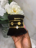 A hand holding a small black velvet jewelry display stand featuring three gold-toned bracelets inspired by Van Cleef & Arpels' Alhambra collection, including a bangle with flower motifs and a cross motif, with white flowers in the background.