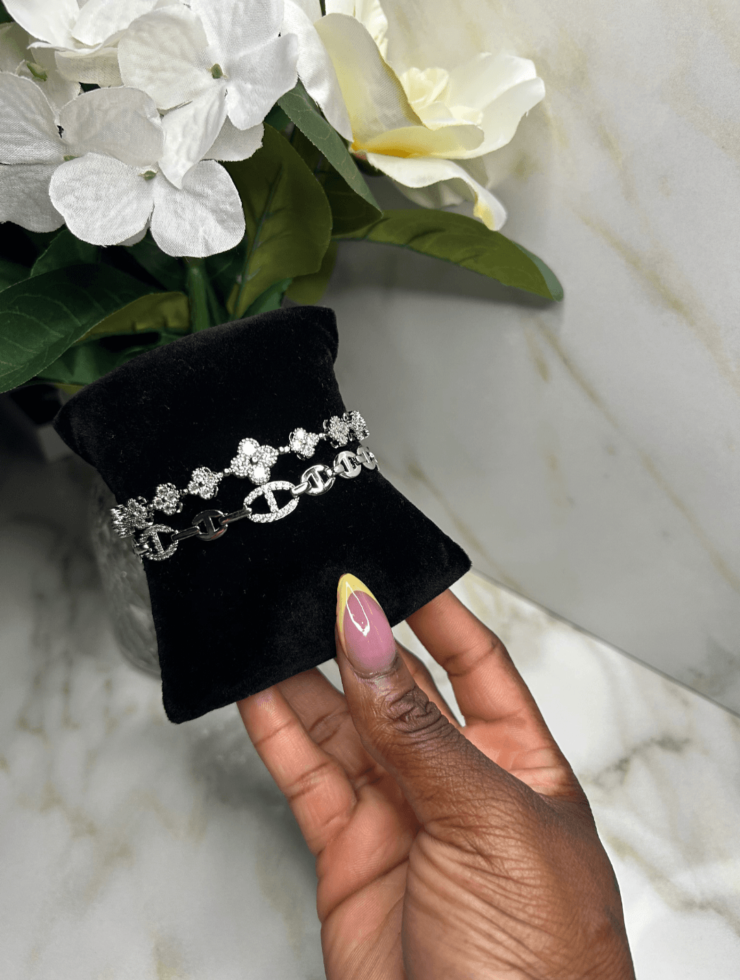 A silver-toned bracelet with interlocking double-C logos and clear stones, displayed on a black velvet jewelry stand held by a hand against a marble surface with white flowers in the background.
